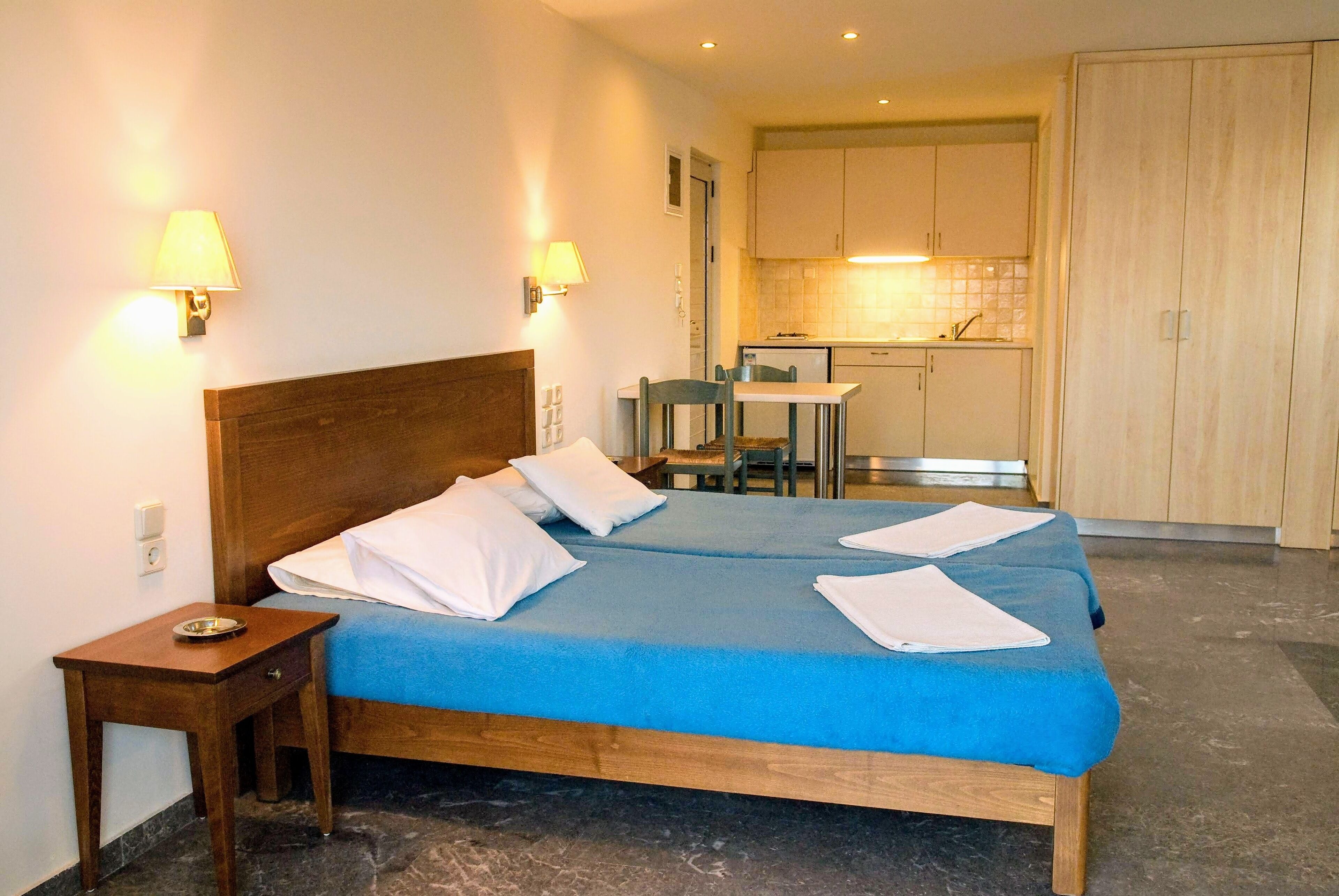 A bedroom with a wooden bed, two bedside tables, a small bedside table with a bowl, a blue bedspread, and a kitchenette in the background.