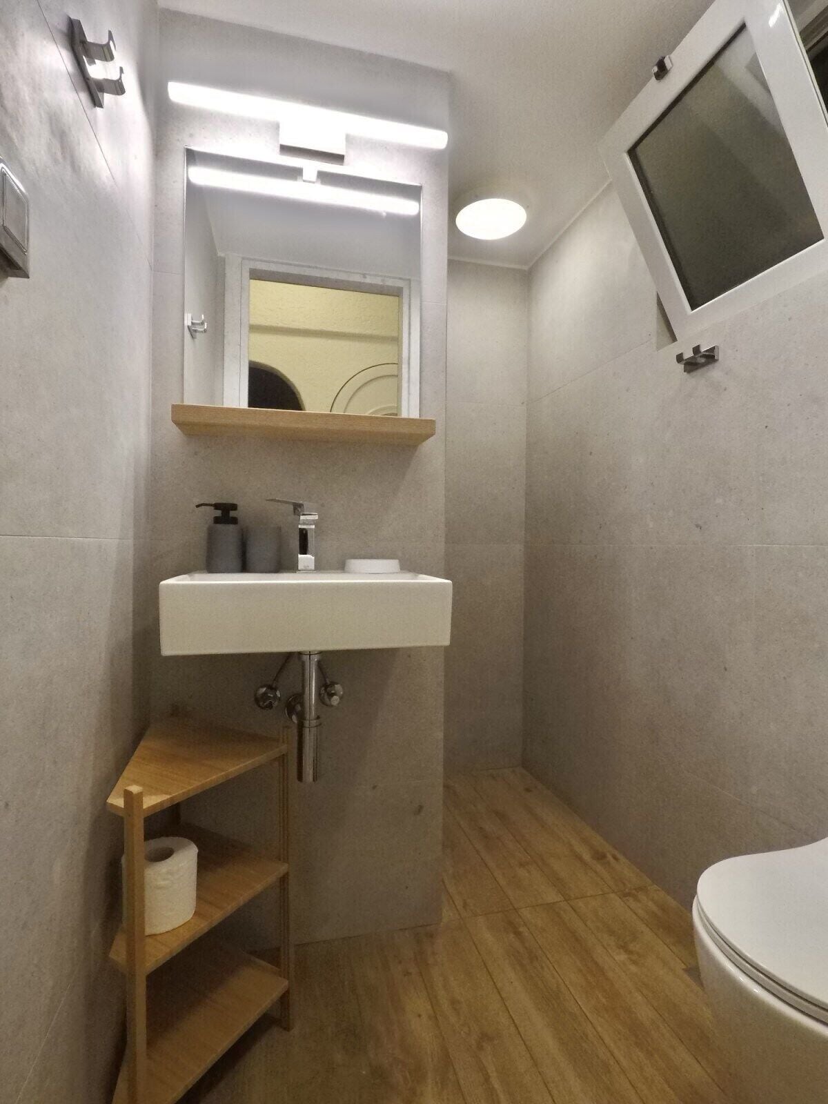 Un baño pequeño con un lavamanos blanco, un espejo, un inodoro y un estante de madera con un rollo de papel higiénico.