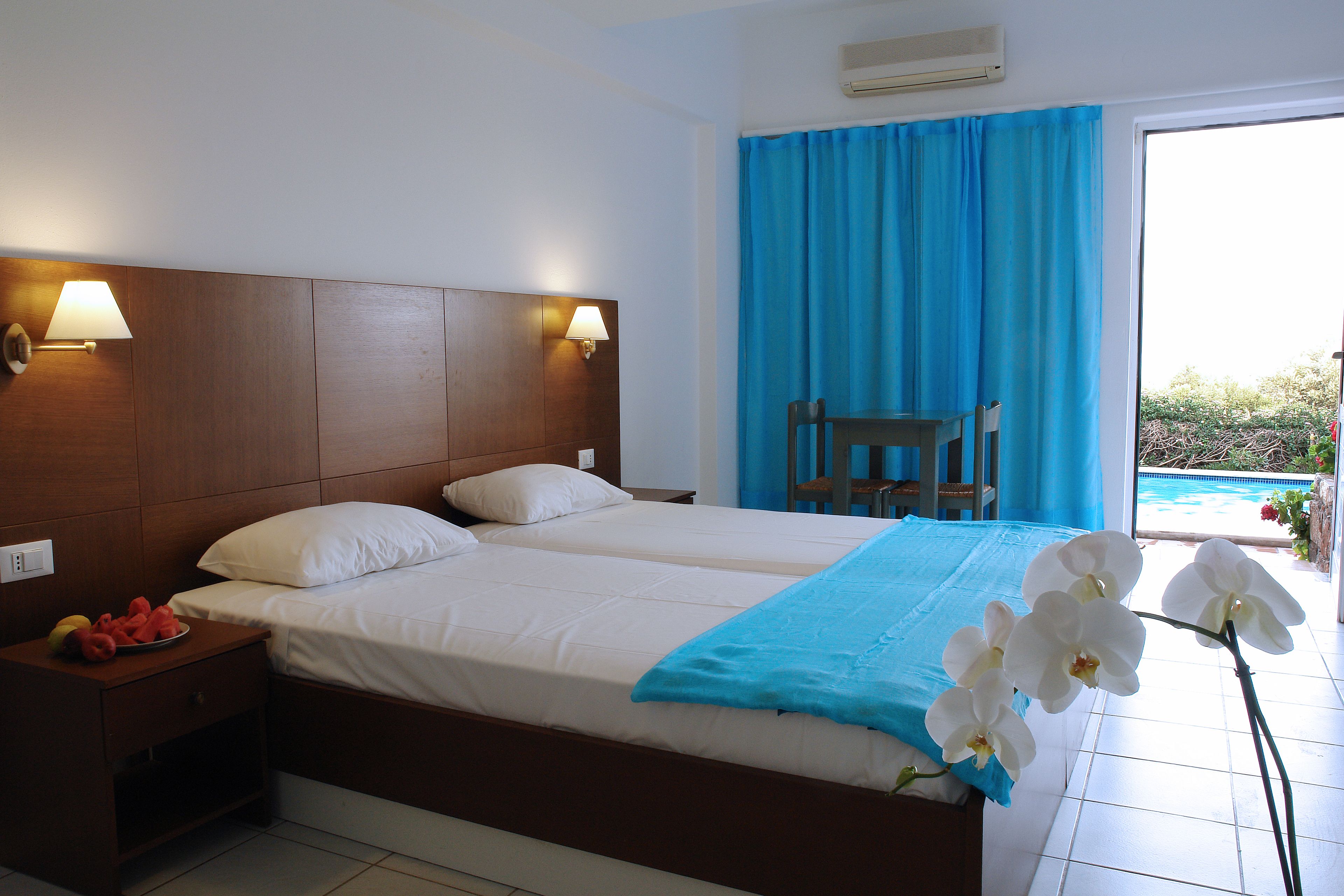 Quarto de hotel com uma cama grande, duas luminárias de cabeceira, uma mesinha de cabeceira com frutas e vista para uma piscina através de uma porta deslizante.