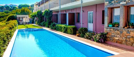 A swimming pool with a wooden deck, surrounded by a garden and a building with a stone facade.