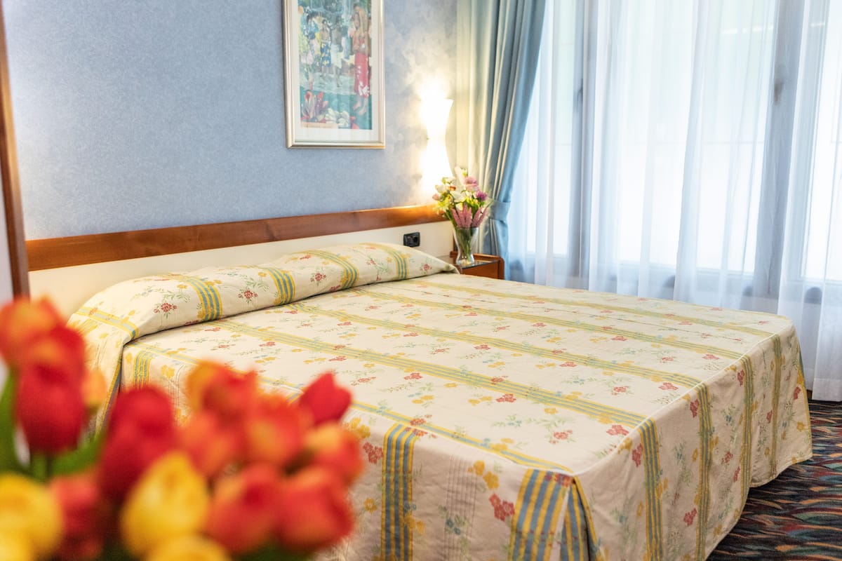 A hotel room with a bed, a nightstand, a vase with flowers, and a painting on the wall.