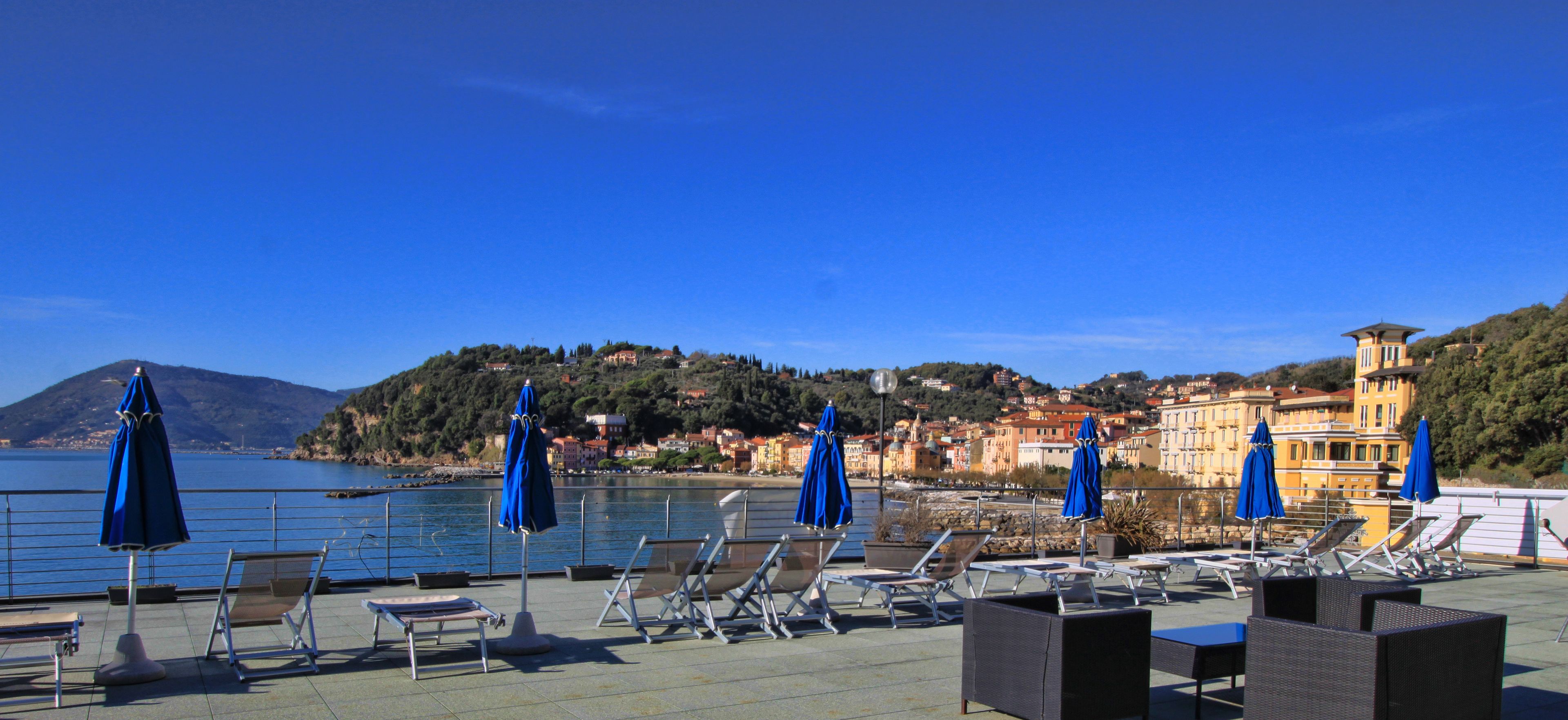 Terraza frente al mar con tumbonas y sombrillas, con vistas a un pueblo costero, edificios y colinas.