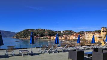 Terraza frente al mar con tumbonas y sombrillas, con vistas a un pueblo costero, edificios y colinas.