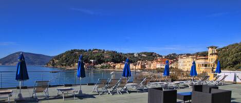 Terraza frente al mar con tumbonas y sombrillas, con vistas a un pueblo costero, edificios y colinas.