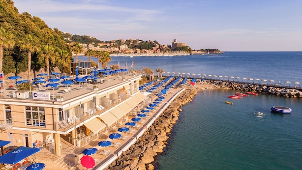 Un hotel frente al mar con mesas y sombrillas al aire libre, una costa rocosa y un muelle que se adentra en el océano.