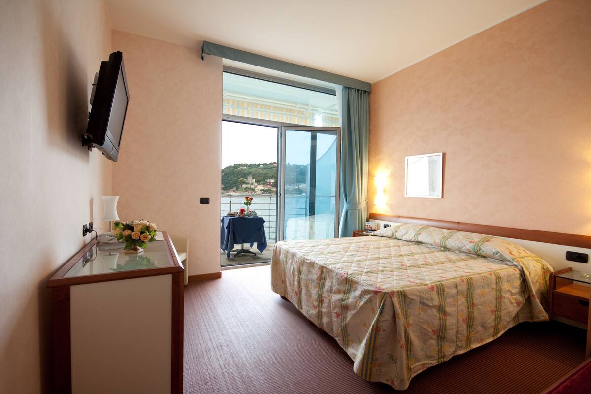 A hotel room with a bed, a desk with a lamp, a TV mounted on the wall, and a view of the outside through a large window.