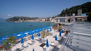 Un complejo turístico frente al mar, con terraza y vistas al océano, que incluye sillas de playa y sombrillas.