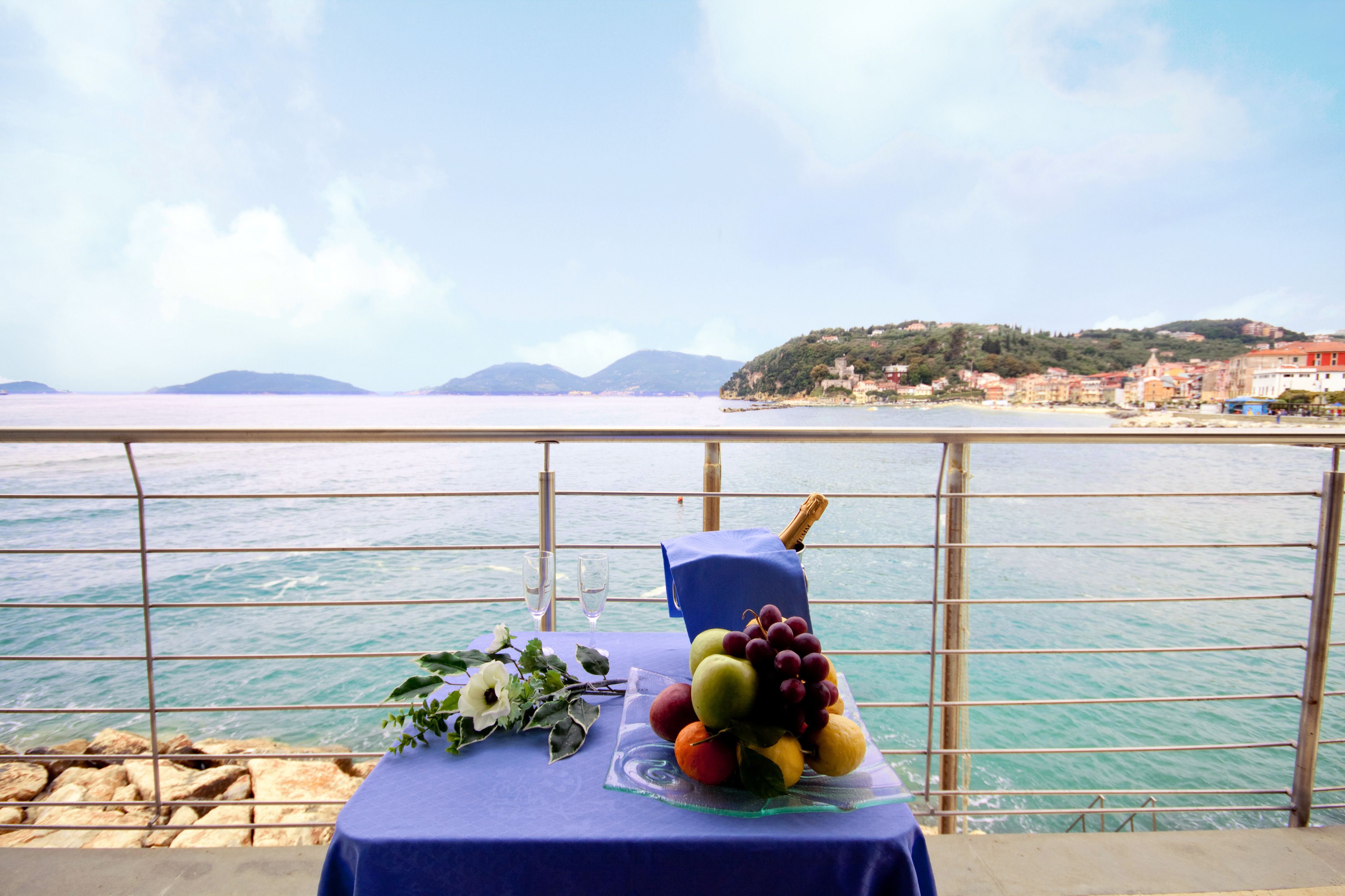A table with a fruit arrangement and a bottle of champagne, overlooking a coastal view with buildings in the background.