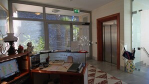 Un mostrador de recepción con computadora, teléfono y diversos útiles de oficina. Hay una ventana con persianas, una puerta con ascensor, y un florero con bolígrafos en el suelo.