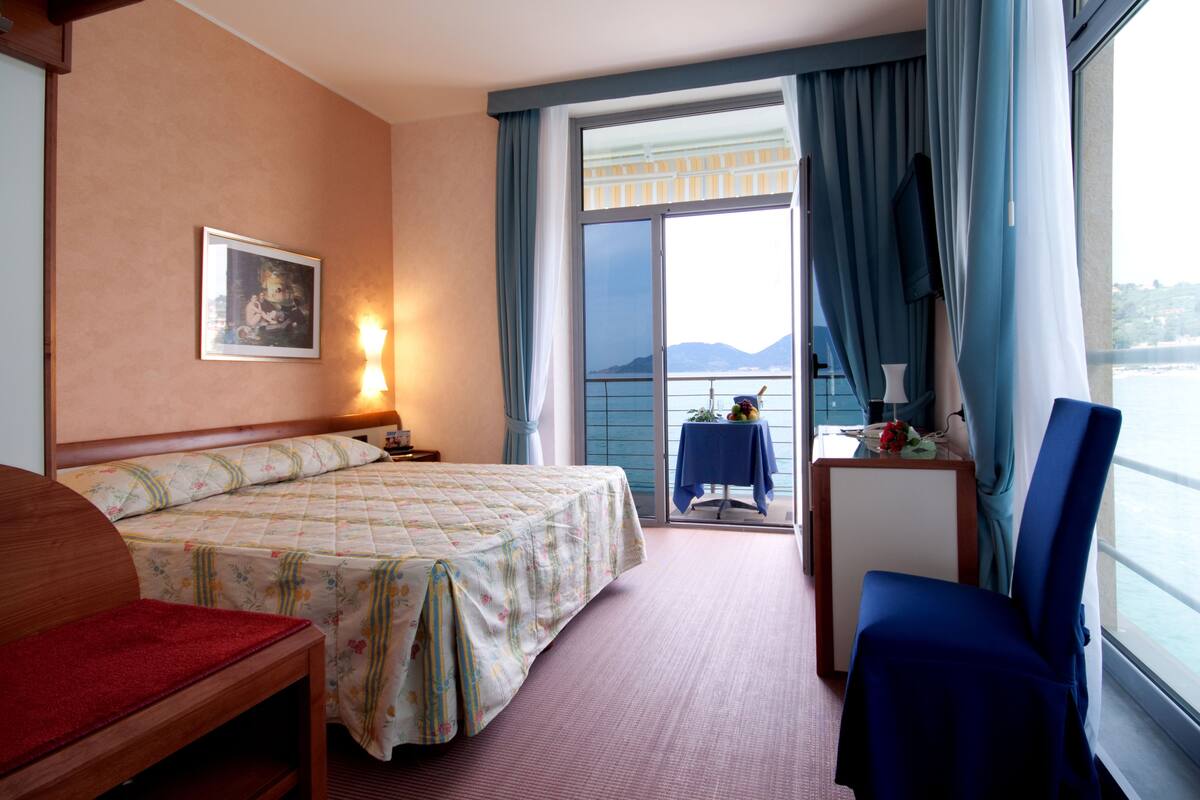 A hotel room with a bed, a desk, a chair, a window with a view of the sea, and a painting on the wall.
