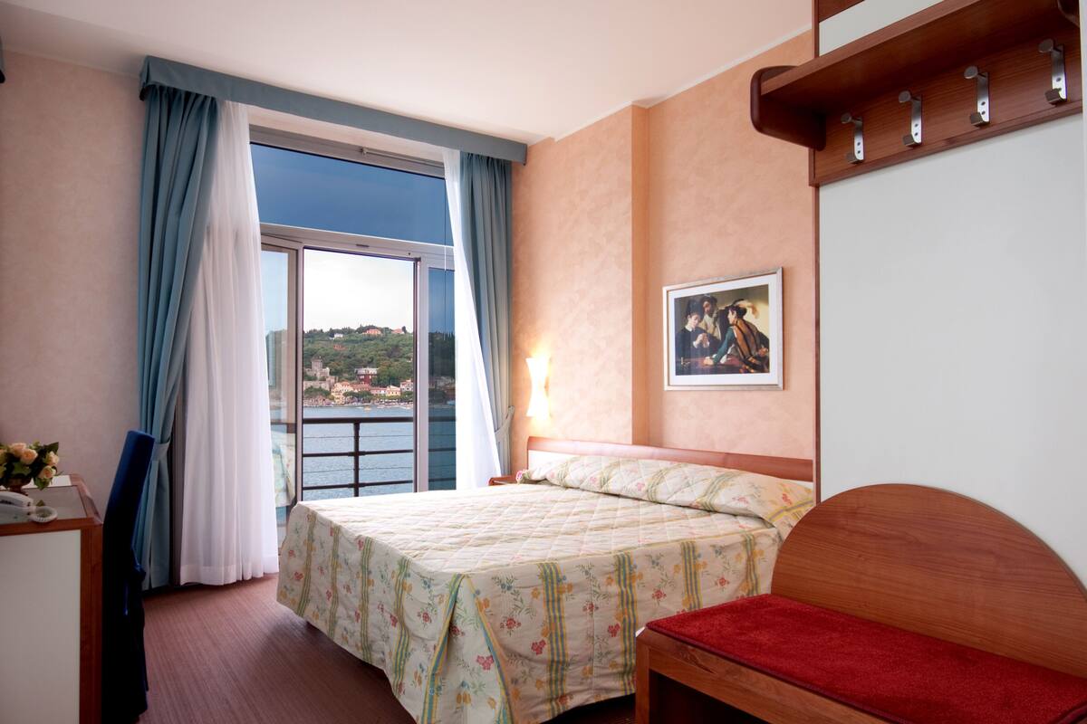 A hotel room with a bed, a desk, a chair, a red bench, a painting on the wall, and a view of the sea through a window.