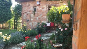 Un jardín empedrado con plantas y flores en macetas, una pequeña mesa y un edificio de ladrillo con una ventana y una planta colgante.
