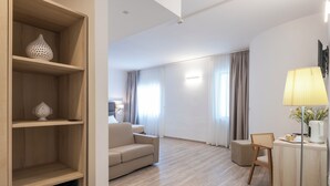 A modern hotel room with a sofa, a small table with a lamp, a wooden cabinet, and a desk with a chair. - Hotel Albània (Otranto)
