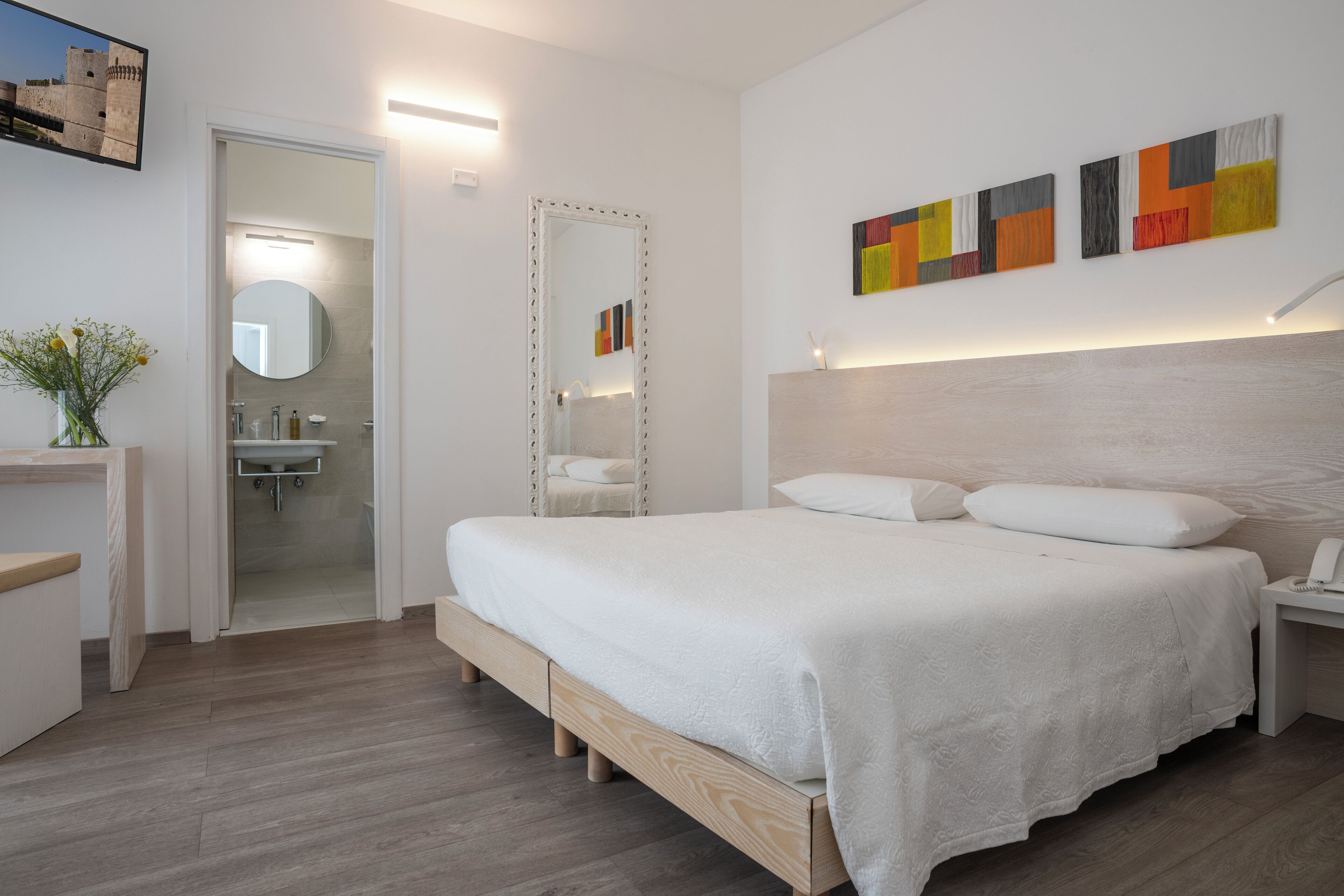 Un dormitorio moderno con cama de madera, una mesita de noche con un teléfono, un espejo y un baño visible a través de una puerta abierta.