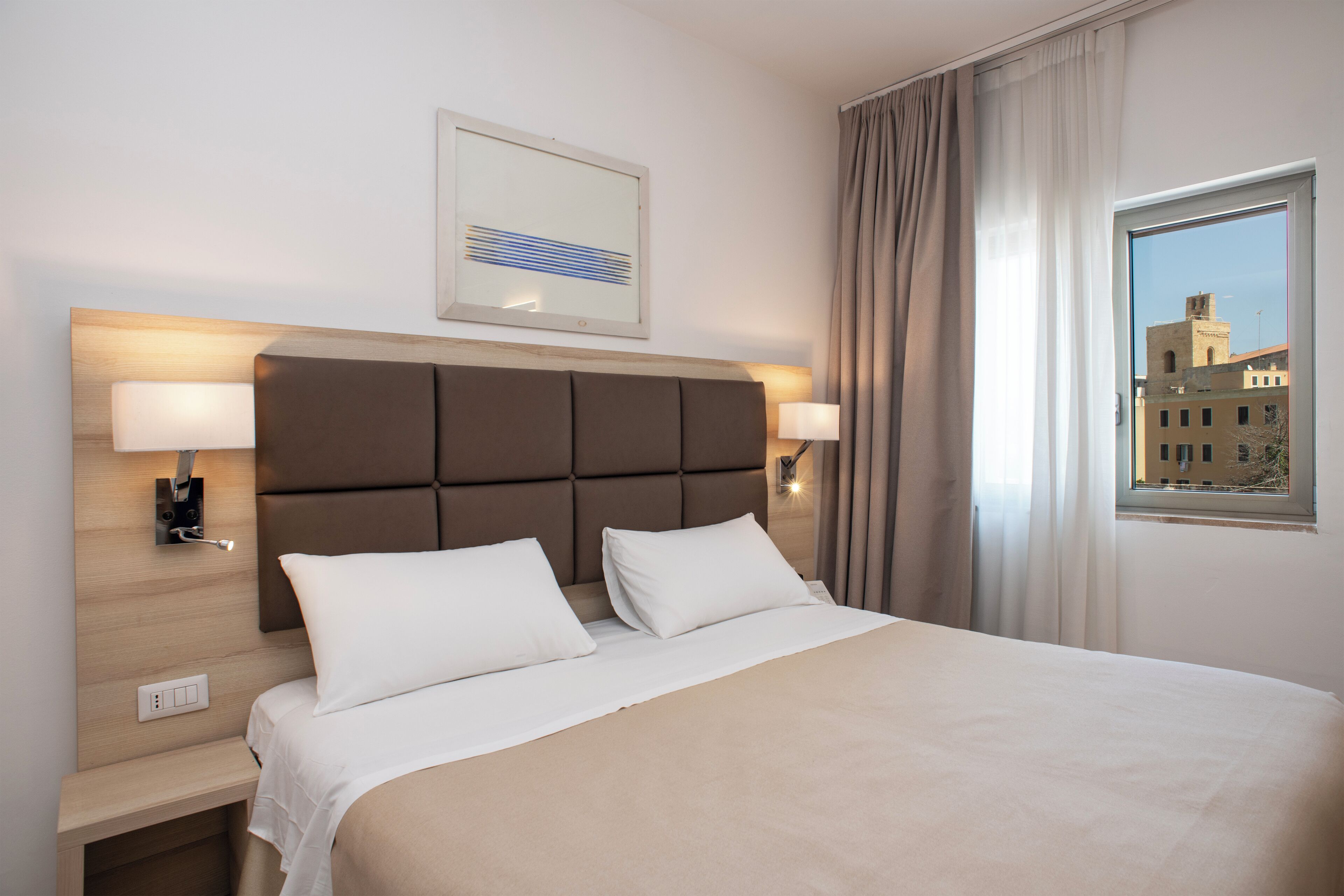 Een hotelkamer met een groot bed, twee bedlampen, een hoofdbord, een ingelijst schilderij, een raam met gordijnen en uitzicht op gebouwen.