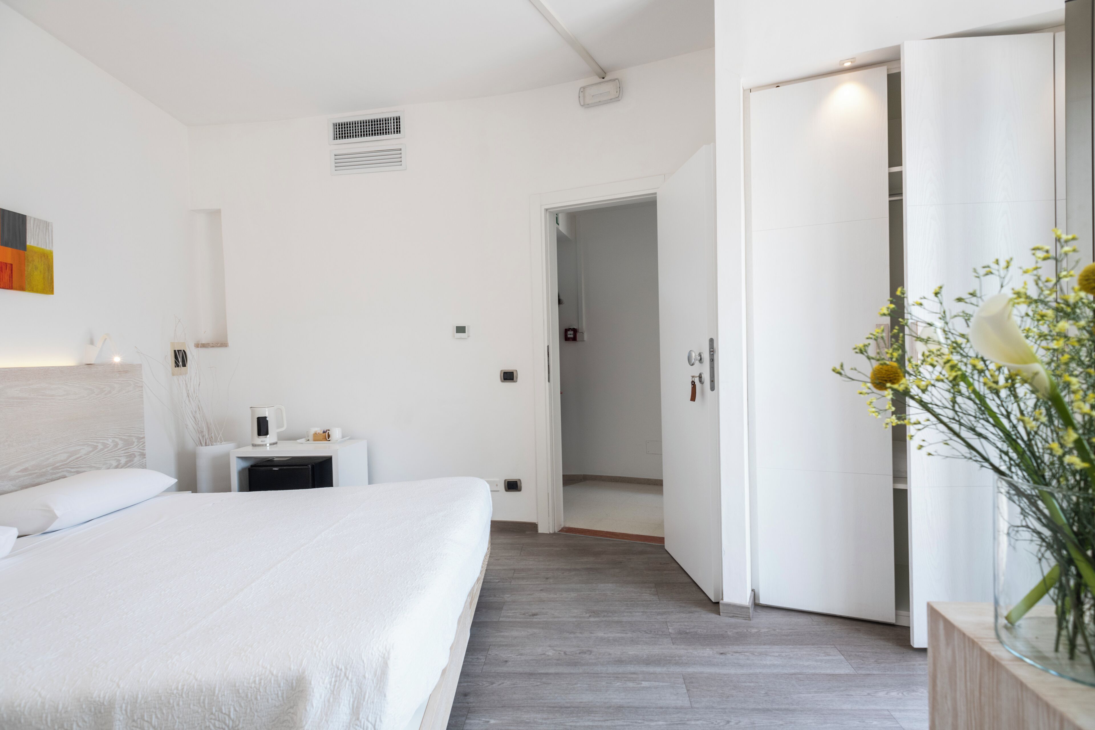 A hotel room with a bed, bedside table, and a small cabinet. There is a door leading to another room.