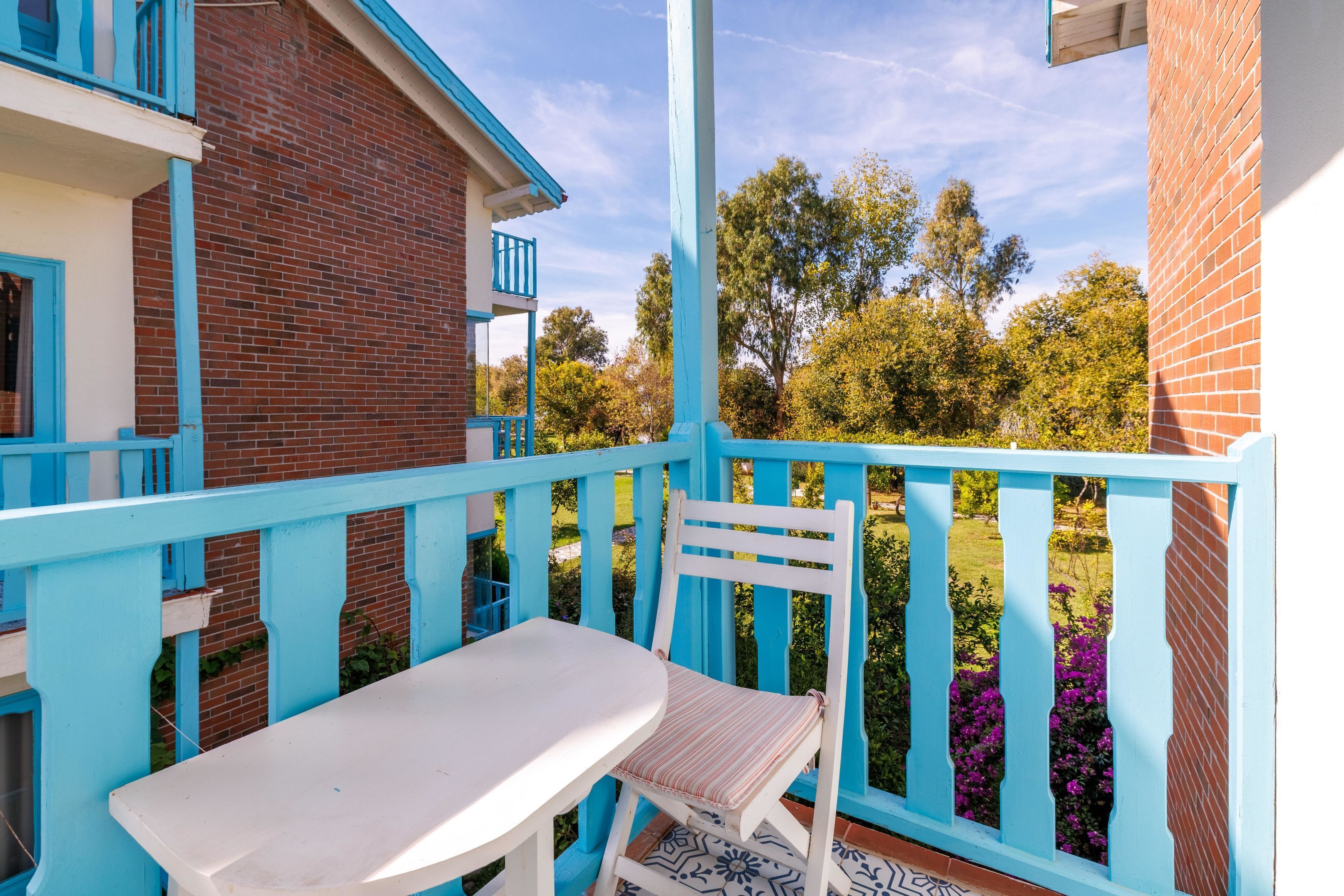 Un balcón con una mesa y una silla, un edificio de ladrillo y vistas a árboles y flores.