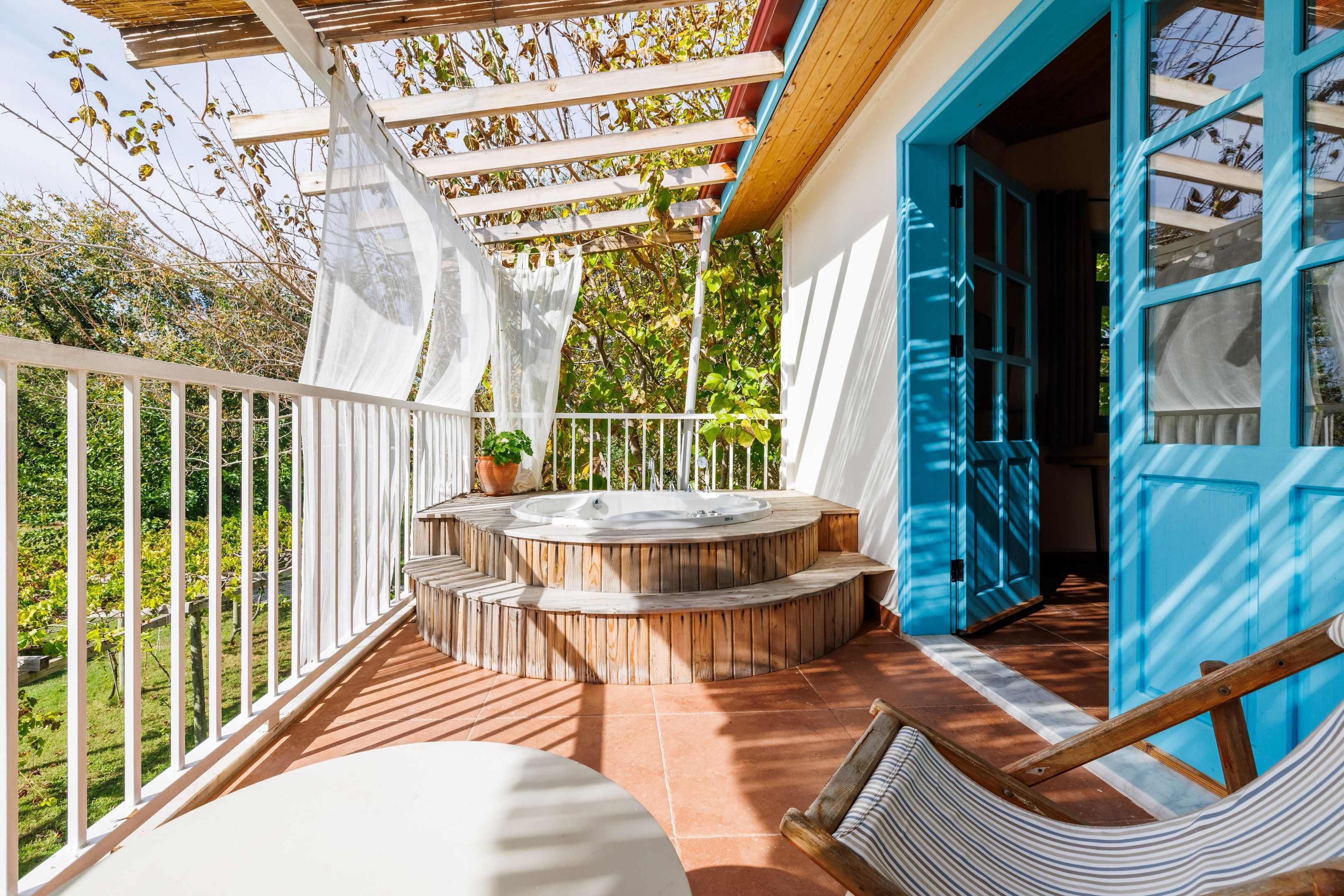 A covered outdoor area with a hot tub, wooden steps, a white railing, and a view of greenery.