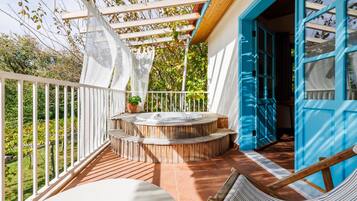 A covered outdoor area with a hot tub, wooden steps, a white railing, and a view of greenery.