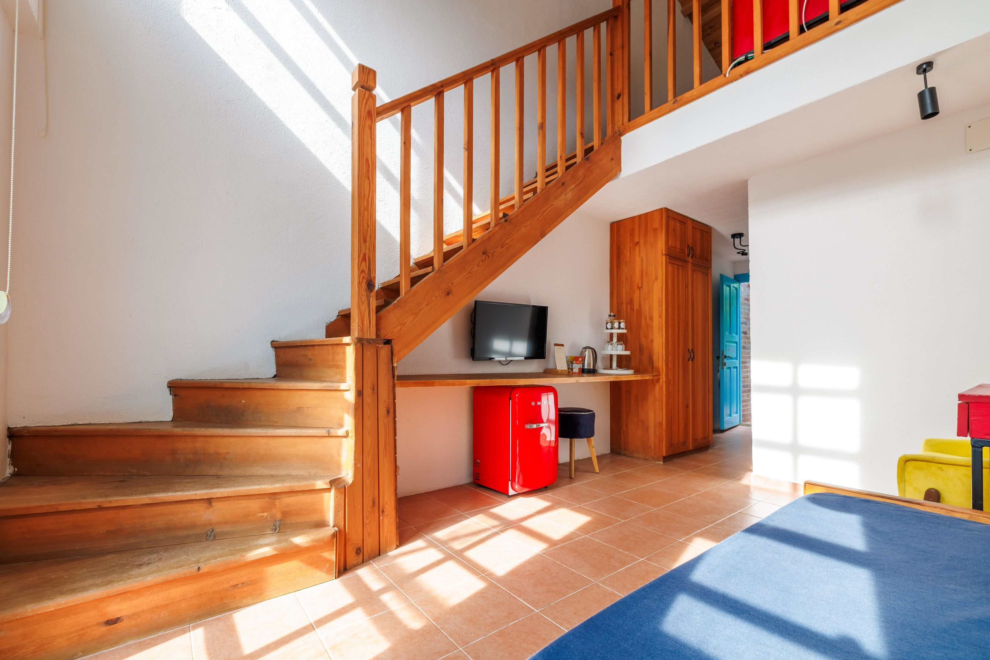 Un escalier en bois mène à une mezzanine où se trouvent un téléviseur, un réfrigérateur rouge et un canapé bleu.