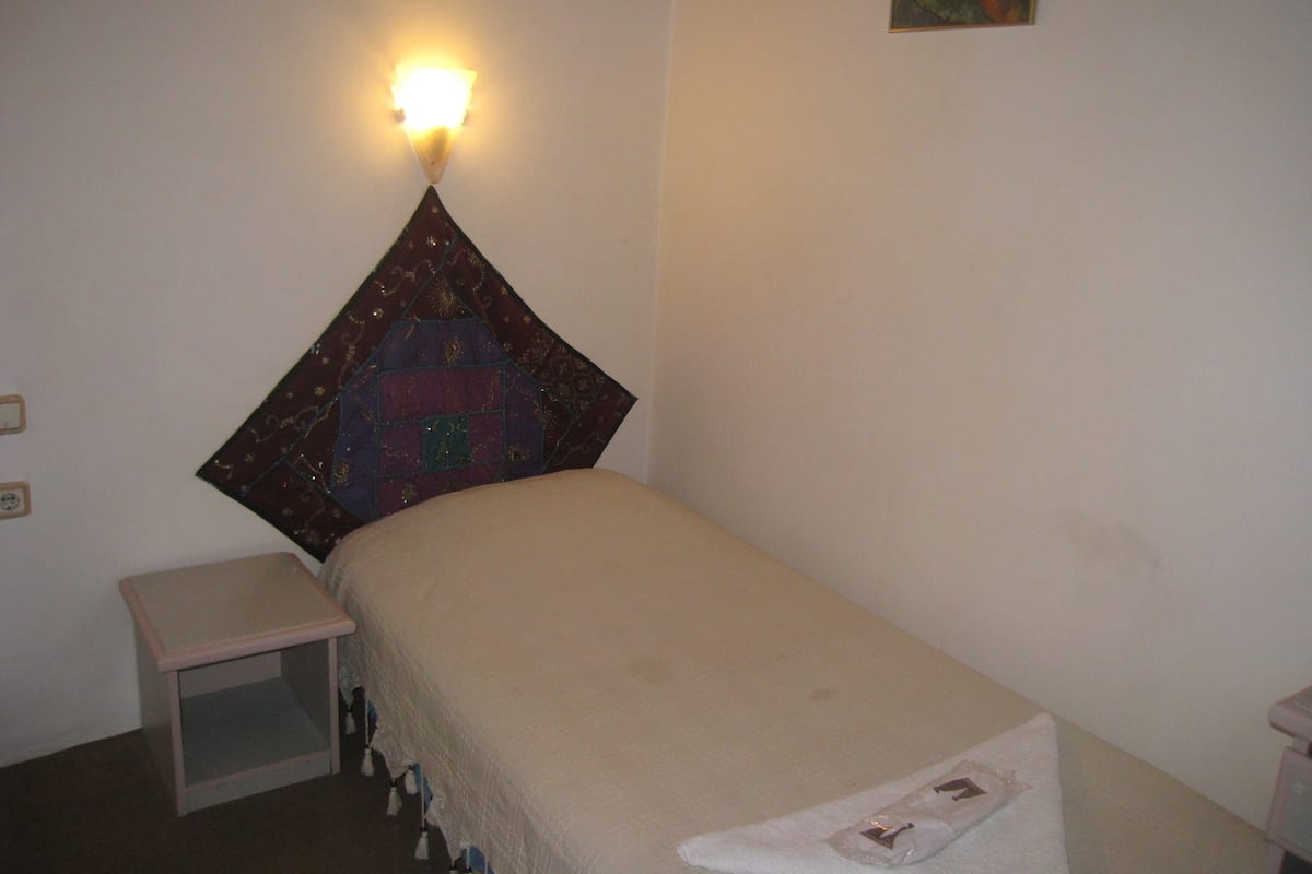 A small, neatly made bed with a white blanket and a decorative pillow. There is a small bedside table and a wall-mounted light fixture.