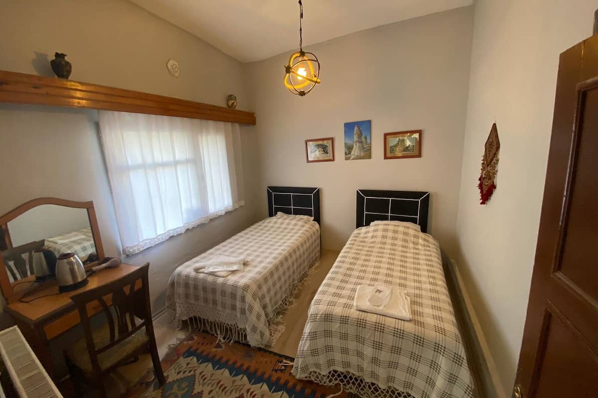 A bedroom with two beds, a desk, a chair, a radiator, a window with curtains, and a hanging light fixture.