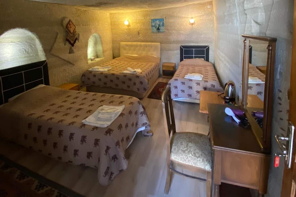 A small, cozy room with two beds, a desk, and a chair. There are wall-mounted lamps and a small rug on the floor.