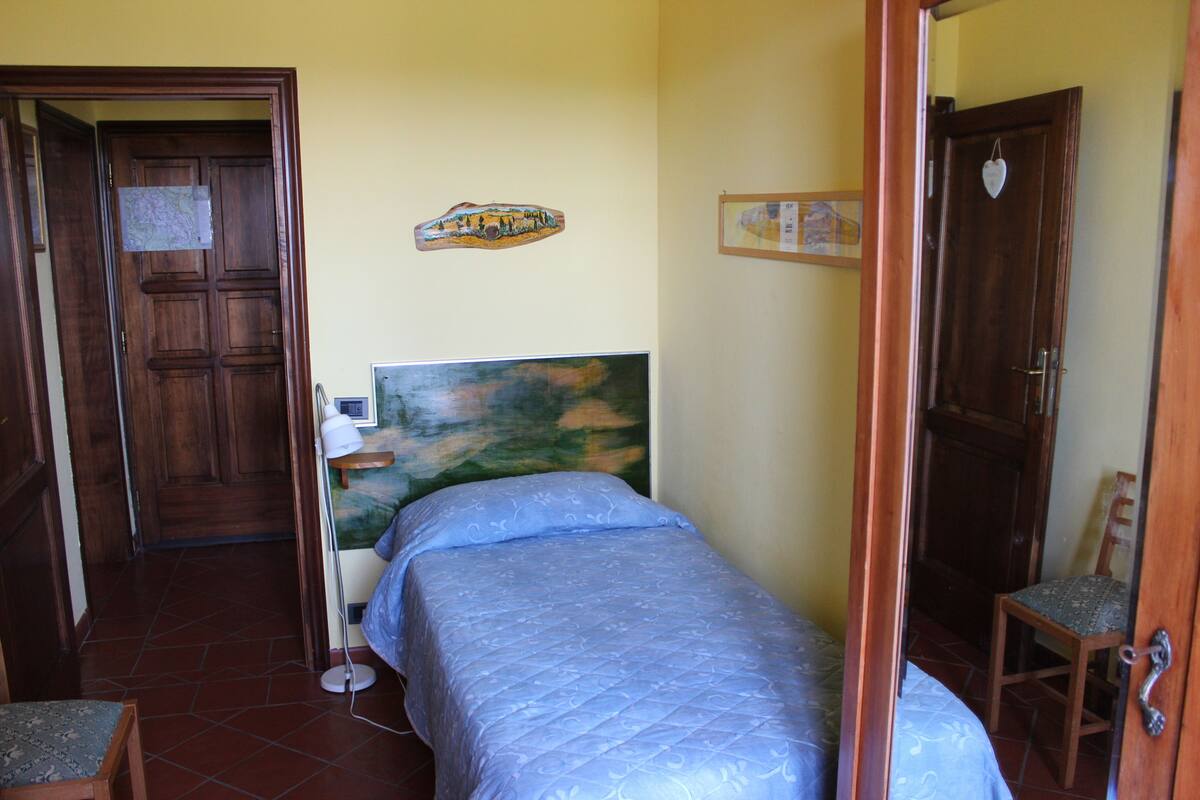 Ein Schlafzimmer mit Bett, Schreibtisch und Stuhl. Eine Holztür und eine Wand mit einem Gemälde sind ebenfalls zu sehen.