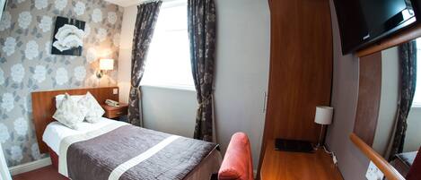 A hotel room with a bed, bedside table, lamp, TV, and a window with curtains.
