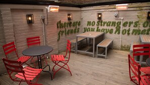 An outdoor seating area with red chairs and tables, a brick wall with green text, and a wooden floor. - Thomas James Hotel (Royal Leamington Spa)
