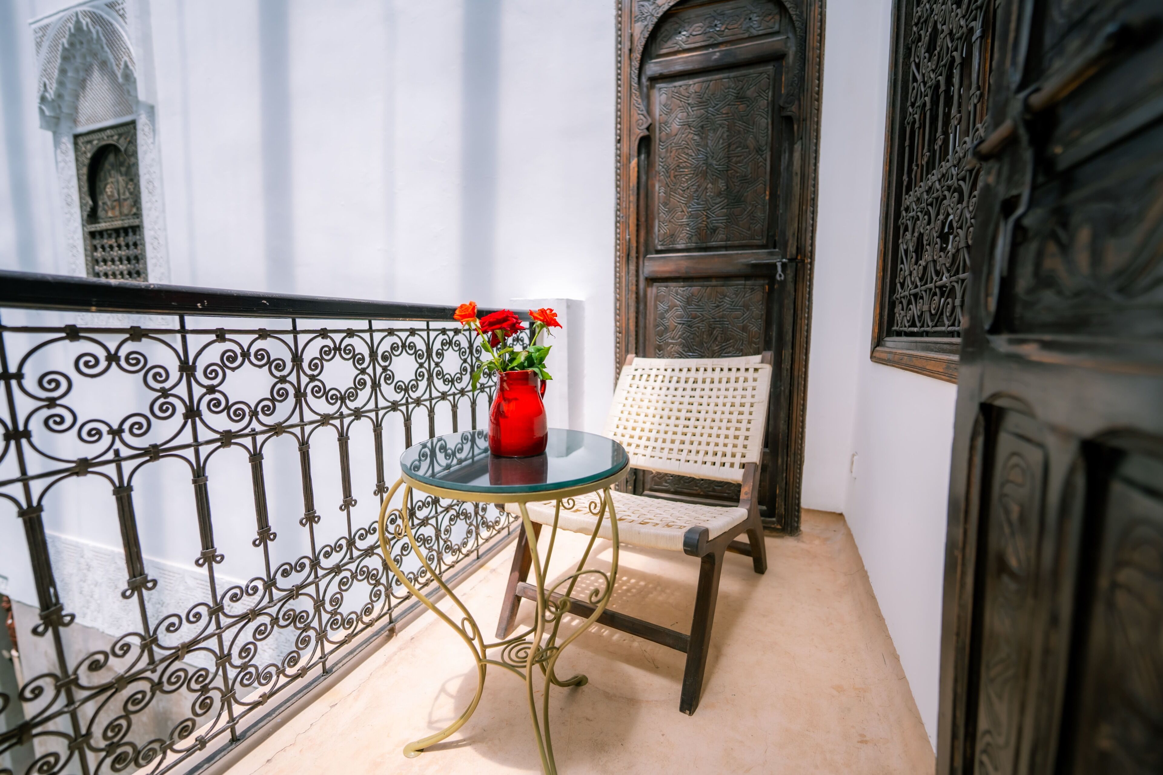 A balcony with a glass table, a red vase with flowers, a white chair, and ornate metal railings.