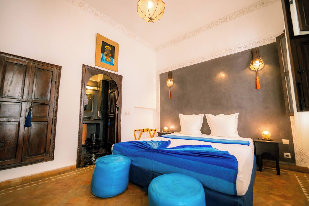 A bedroom with a large bed, two blue ottomans, a wooden wardrobe, and a framed picture on the wall.