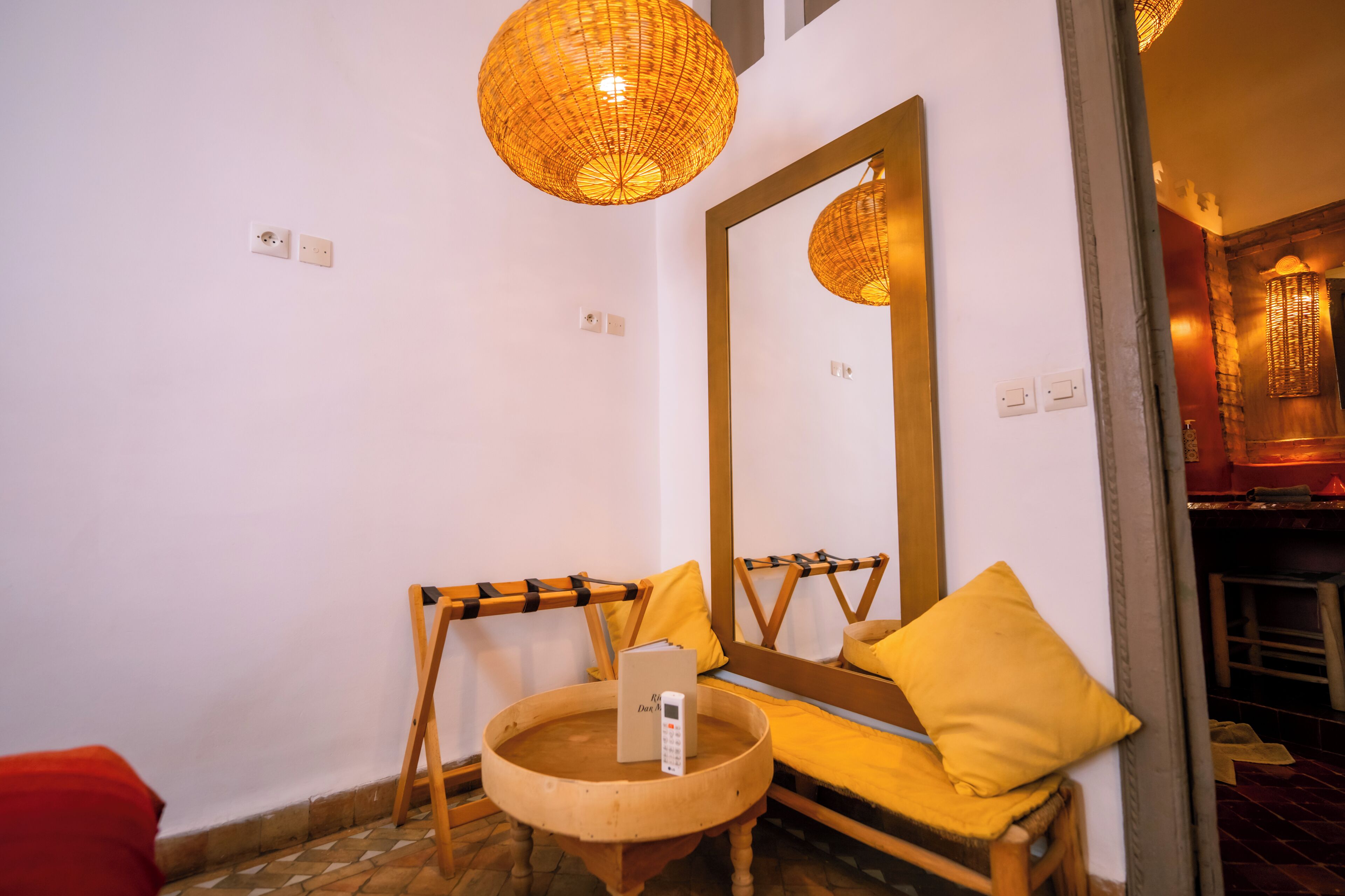 A bathroom with a large mirror, a wooden chair, a small round table with a remote control, and a yellow pillow.