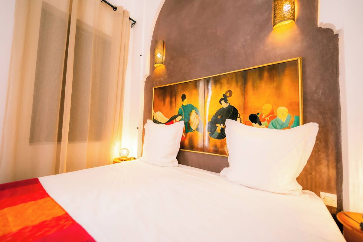 A hotel room with a large bed, white bedding, a painting on the headboard, and a nightstand with a lamp.