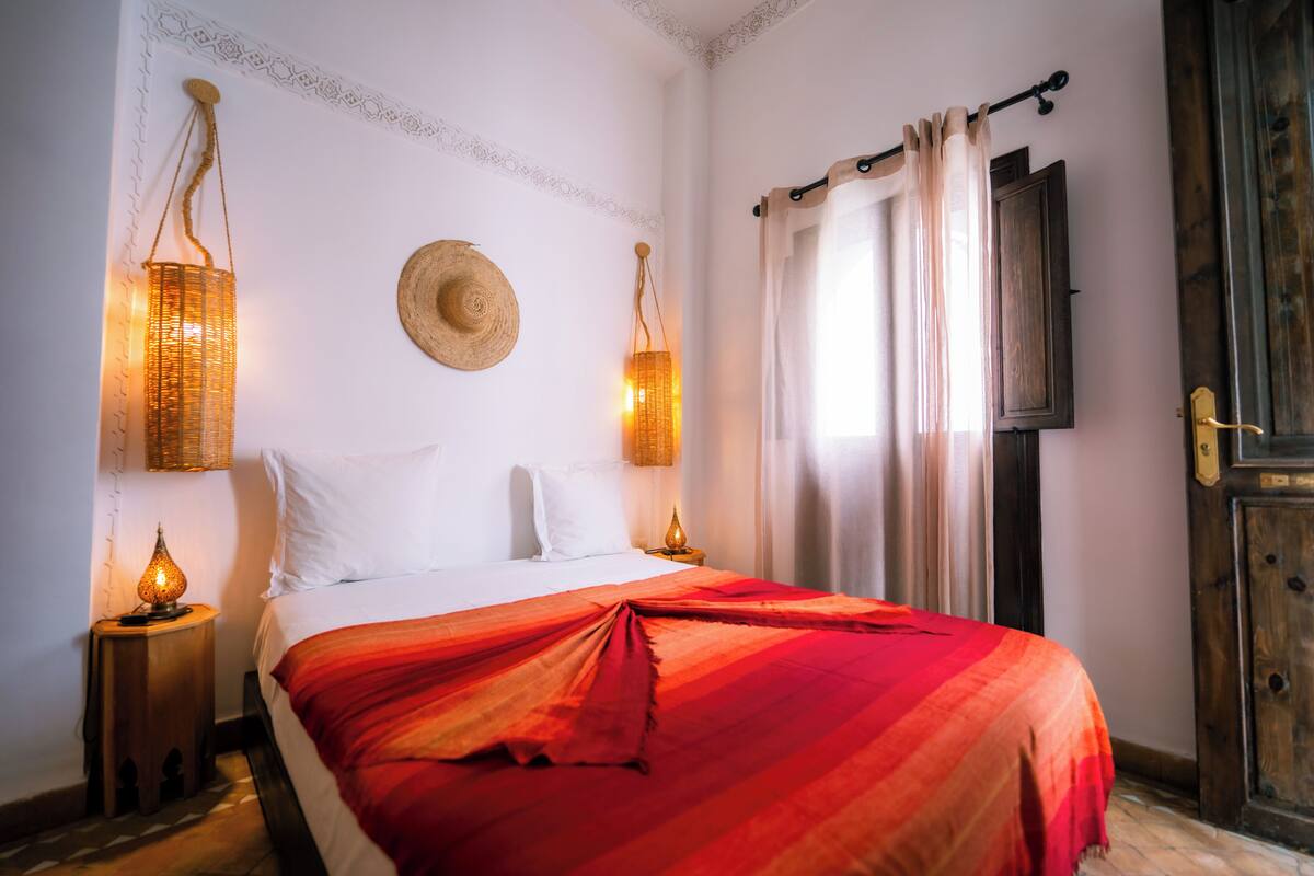 A neatly made bed with a red blanket, two wall-mounted lamps, a straw hat on the wall, and a wooden door.
