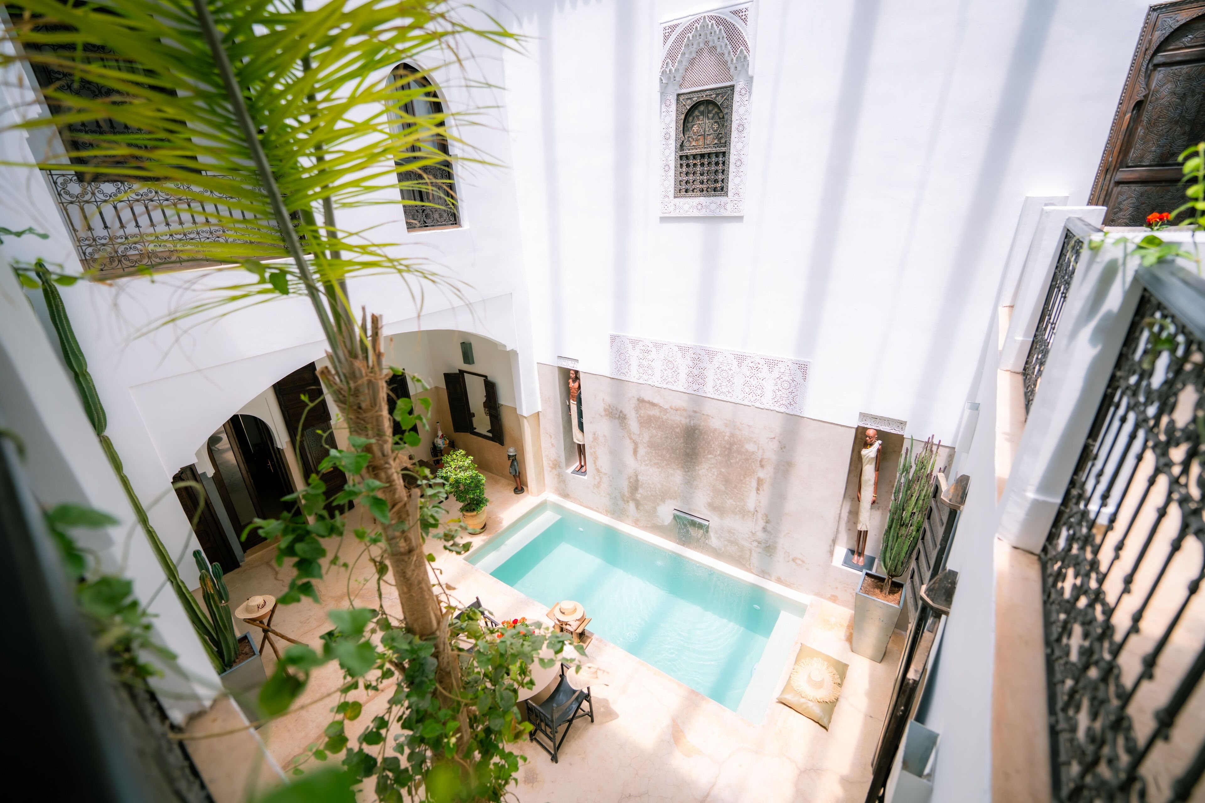 A courtyard with a swimming pool, surrounded by white walls and arches, with potted plants and a small table.