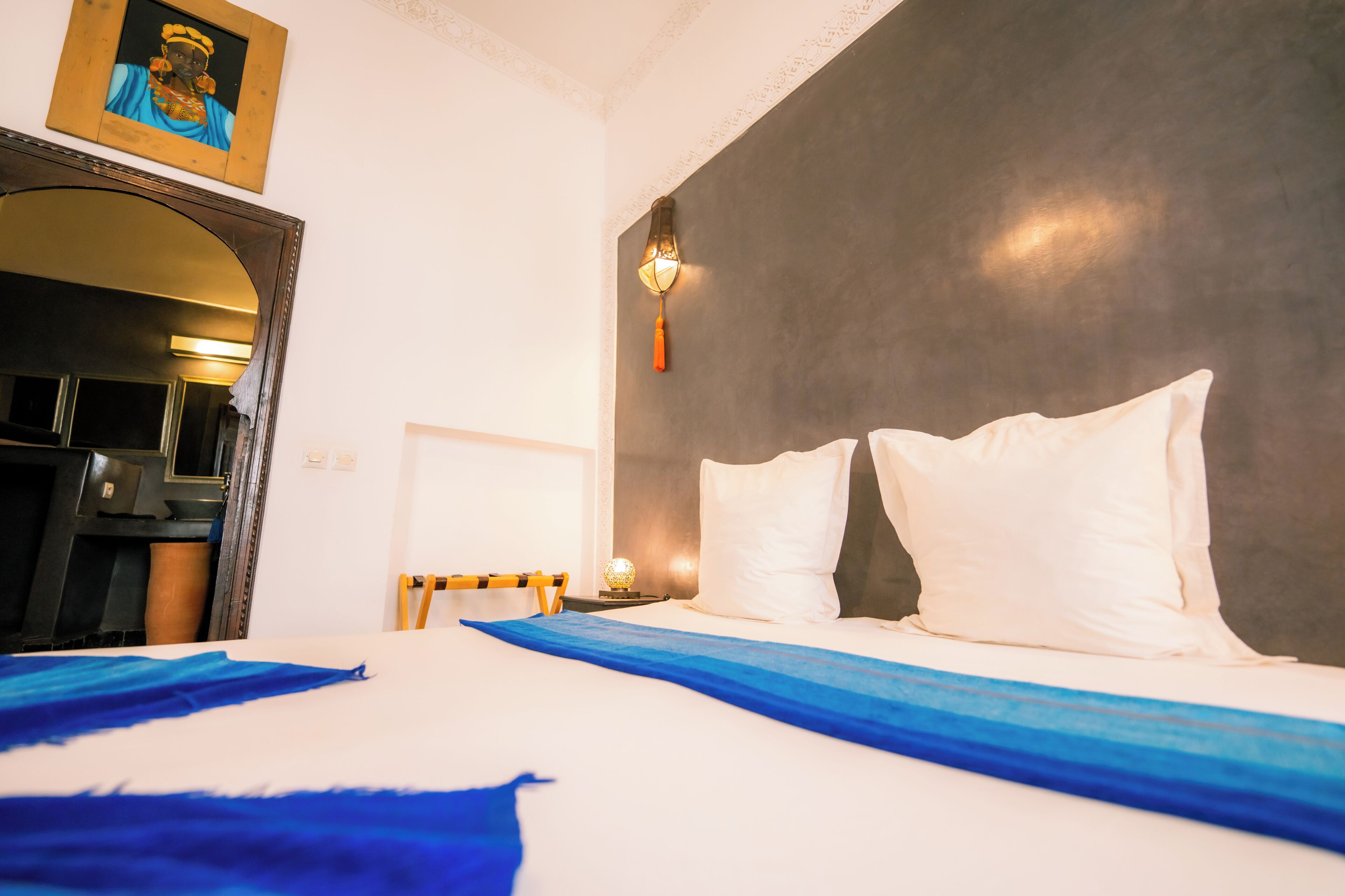 Habitación de hotel con una cama grande, almohadas blancas, colcha azul, una lámpara de pared y un cuadro enmarcado sobre la cama.