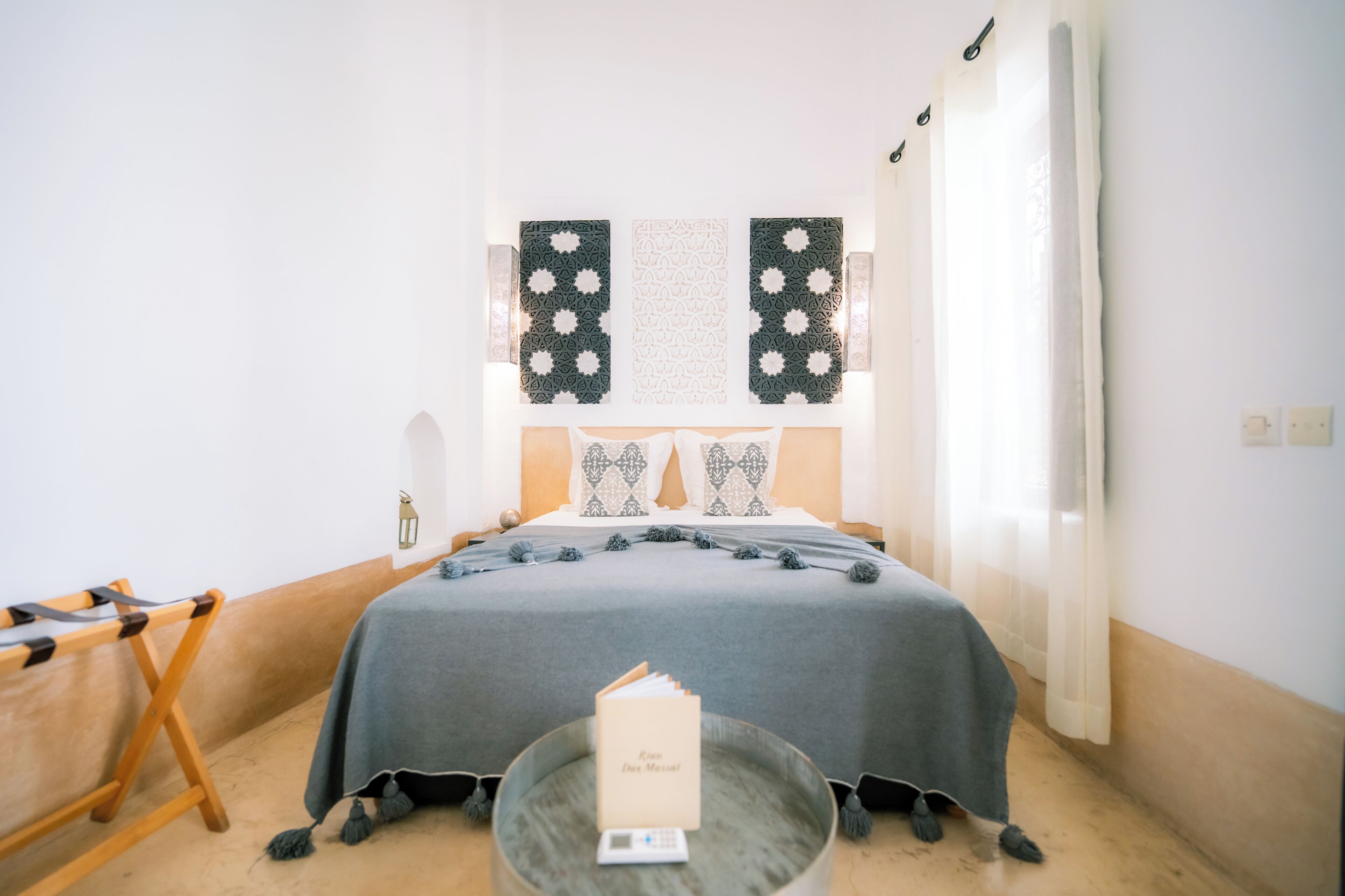 Un dormitorio ordenado con una cama en el centro, una mesita con una tarjeta y una ventana con cortinas.