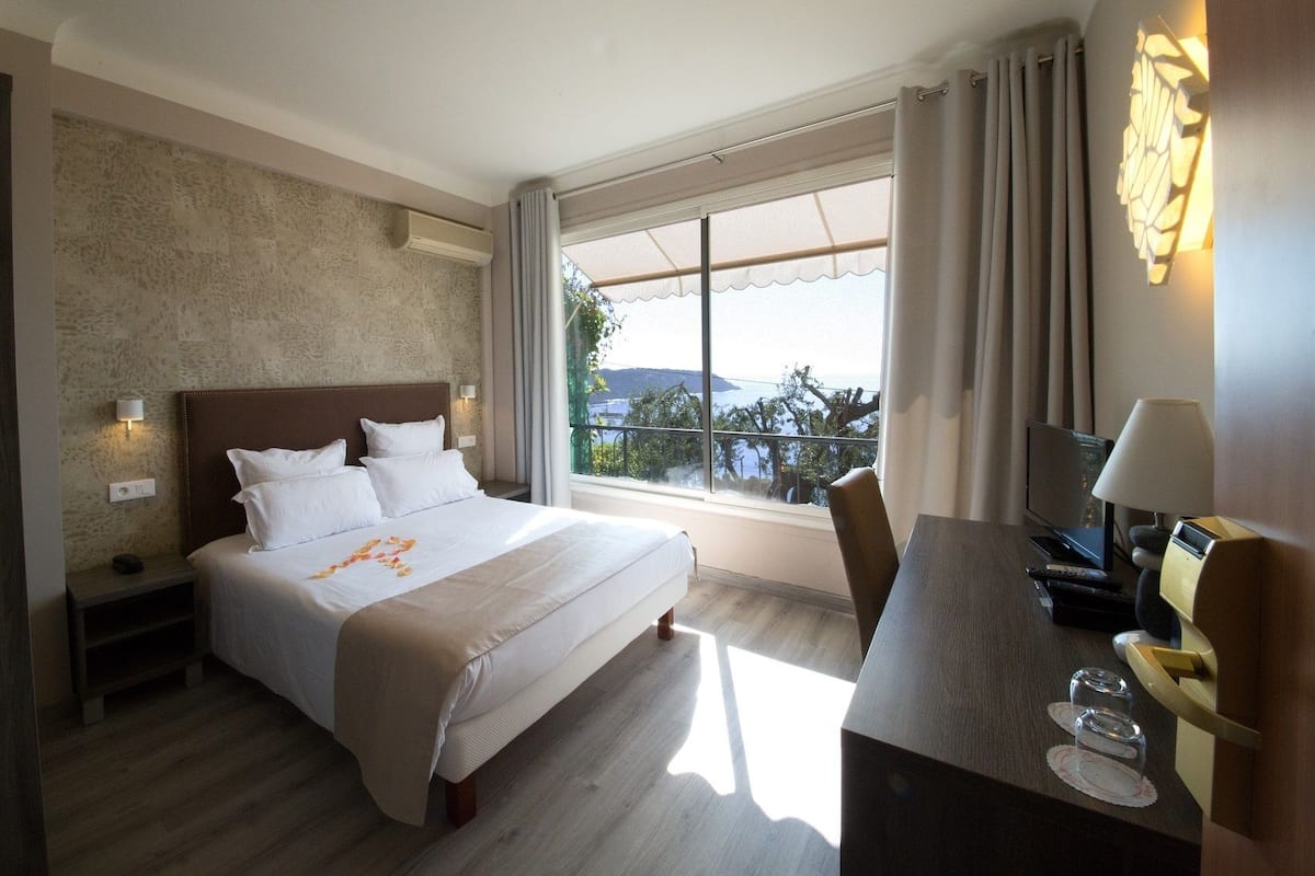 A hotel room with a large bed, a desk with a lamp, a TV, and a view of the outdoors.