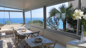 Un patio iluminado por el sol con mesas y sillas de madera, un florero con flores blancas y vista al mar a través de grandes ventanales.