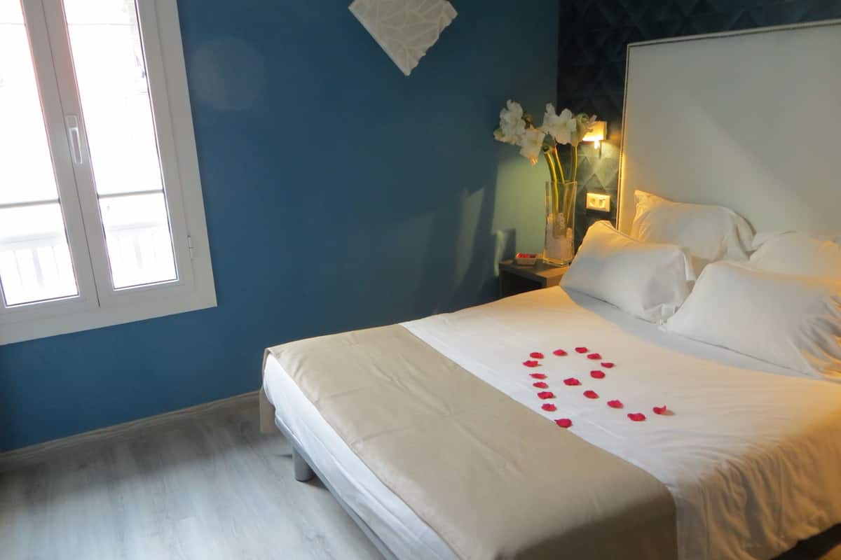 A neatly made bed with white linens and a heart-shaped arrangement of rose petals on the bedspread.