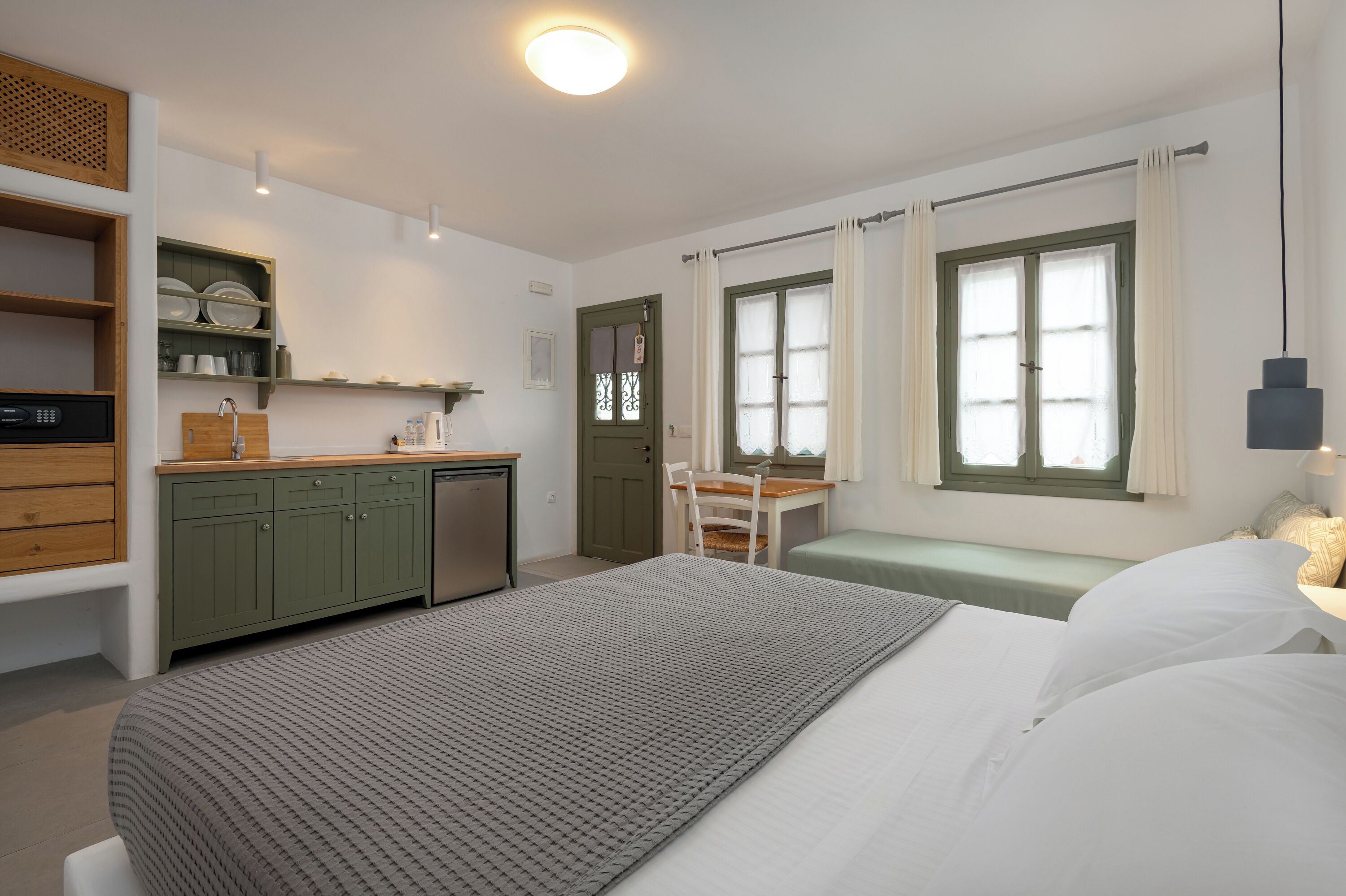A bedroom with a bed, a desk, a chair, a window, and a kitchenette.