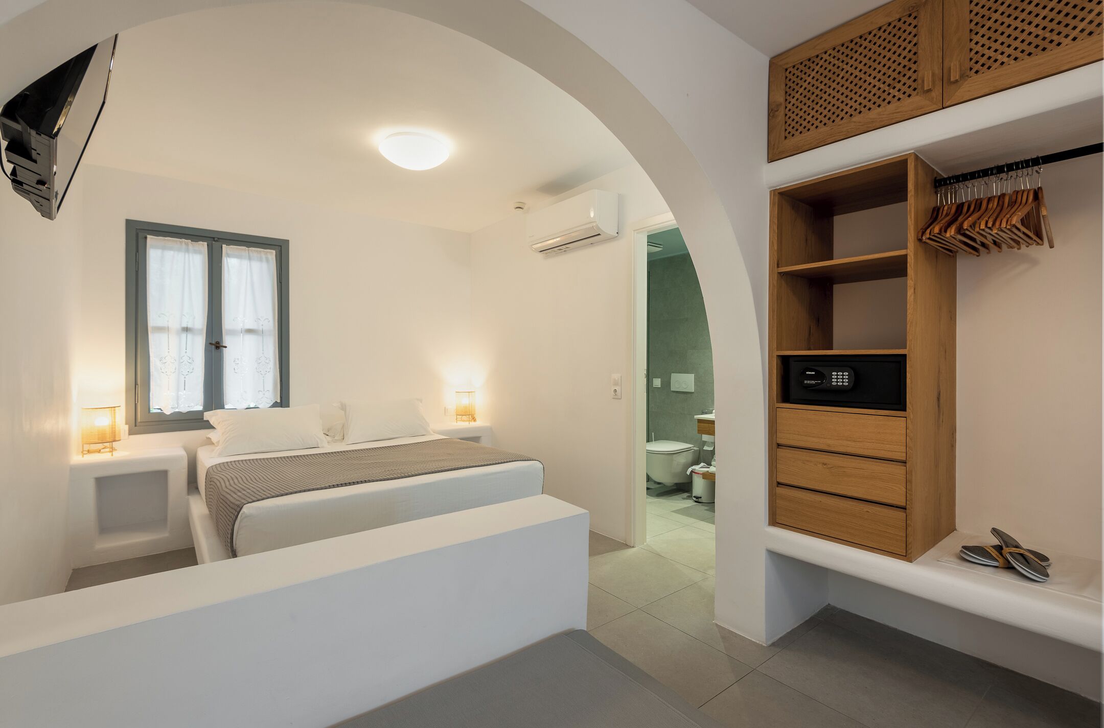 A bedroom with a bed, bedside lamps, a TV, a wardrobe, and a bathroom visible through an archway.