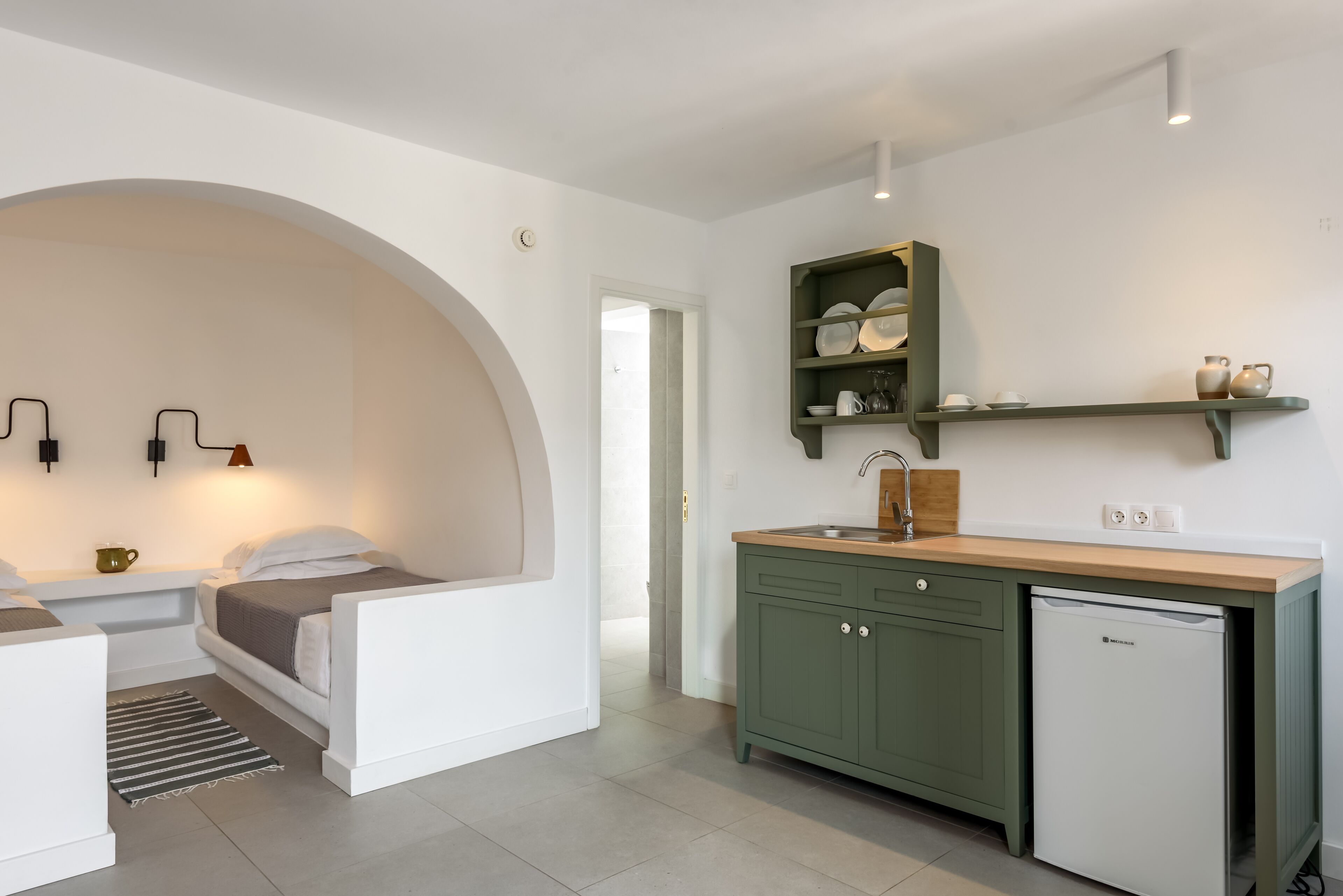 Un dormitorio moderno con un gran ventanal arqueado, una pequeña kitchenette con refrigerador y una mesita de noche con lámpara.