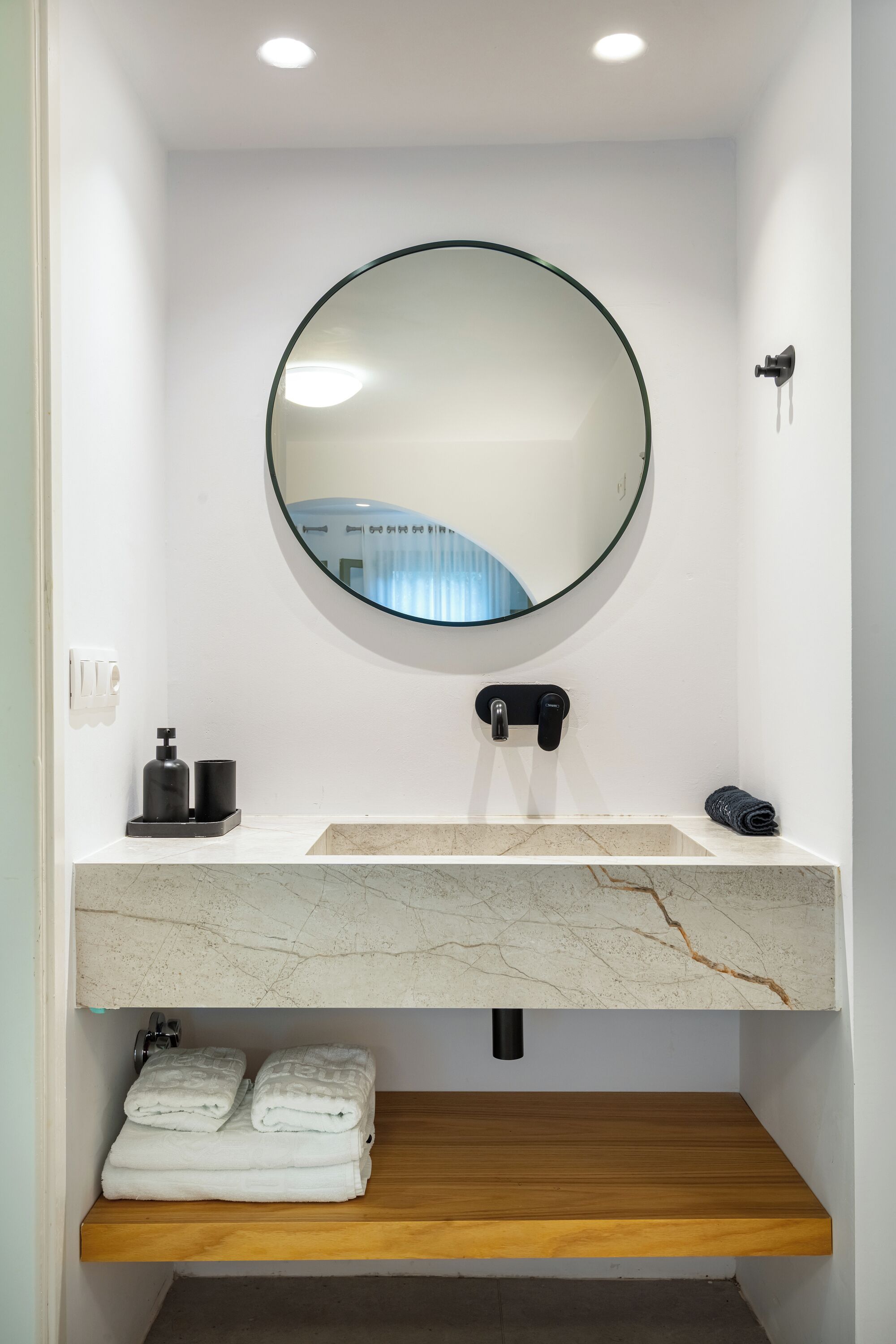 A bathroom with a round mirror, a white sink, a wooden shelf, and a towel rack.