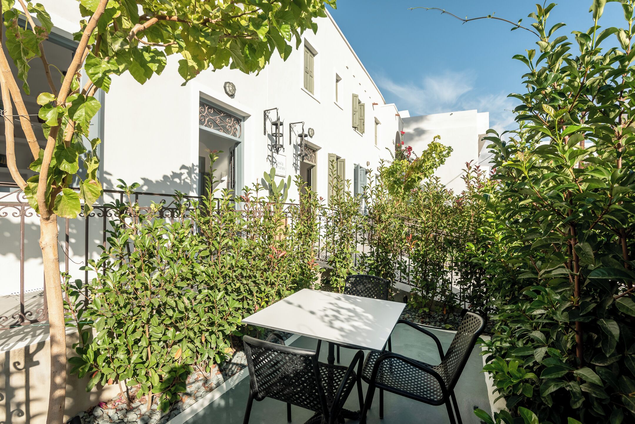 Un bâtiment blanc doté d’un balcon, une table avec des chaises et de la verdure tout autour.