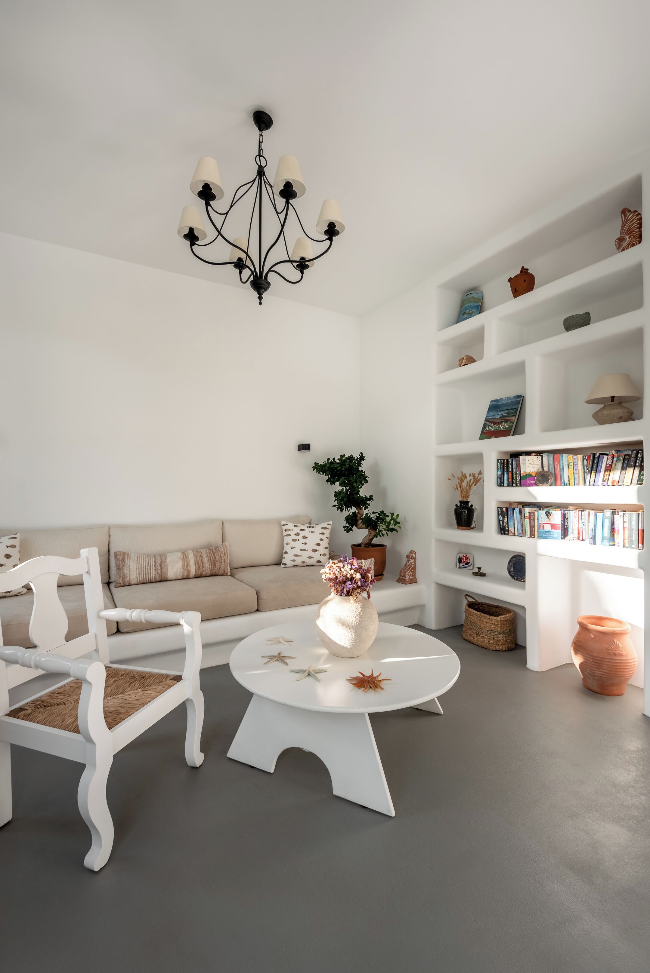 A living room with a white sofa, a white chair, a round coffee table, a black chandelier, and a bookshelf with books and decorative items.