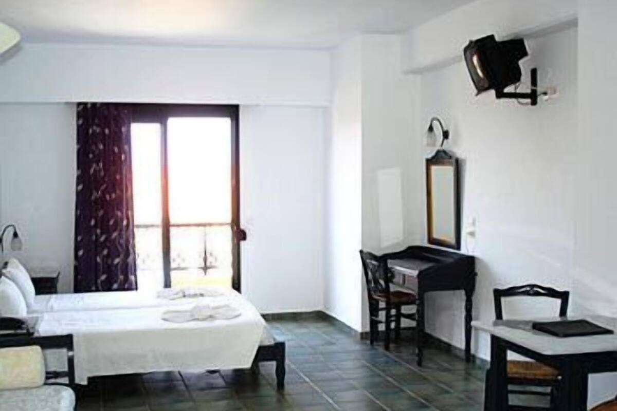 A room with a bed, a desk, and a chair. There is a window with curtains, a wall-mounted lamp, and a mirror.