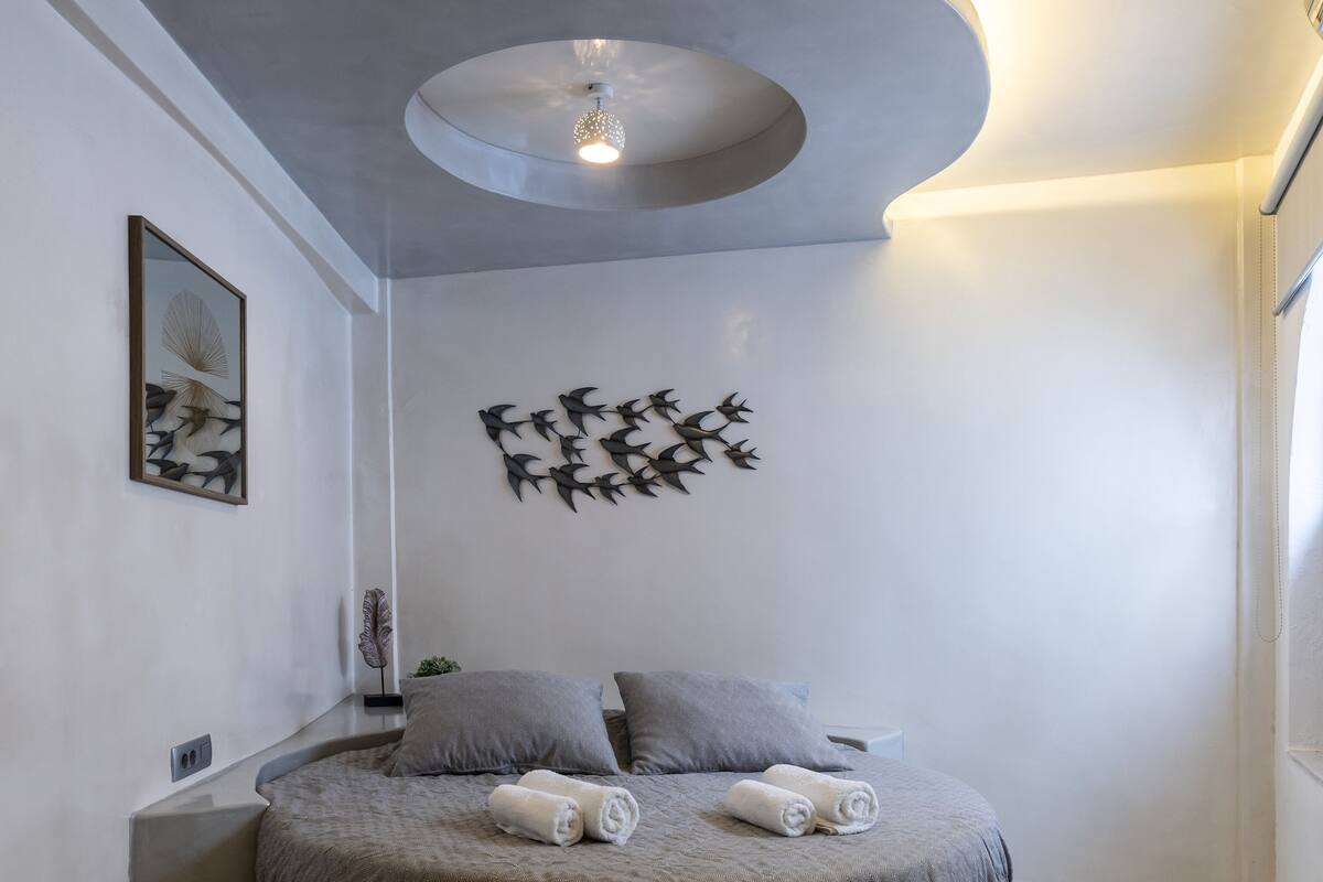 Un dormitorio moderno con cama redonda, ropa de cama gris, una obra de arte en la pared y un diseño de techo original.