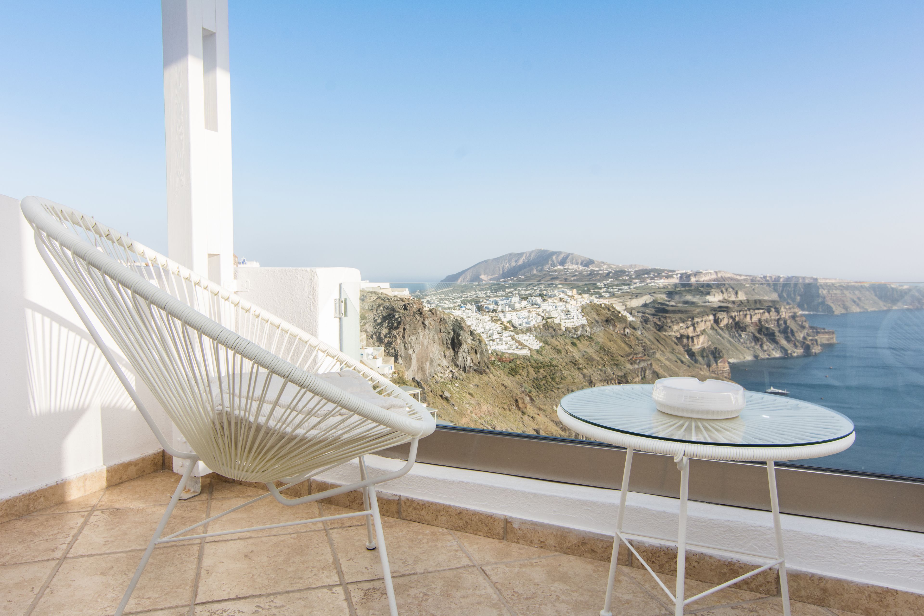 Un balcón con una silla y una mesa blancas, con vistas a un paisaje costero con acantilados y el mar.